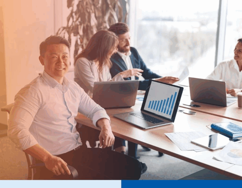 asian man in a corporate suit attire smiling with paperwork and laptop in office full of people