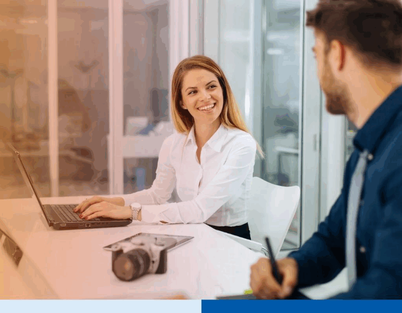 Two professionals collaborating in a modern office, smiling while discussing work on laptops 