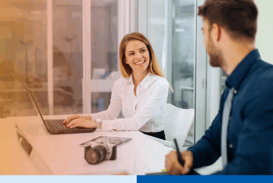 Two professionals collaborating in a modern office, smiling while discussing work on laptopsÂ